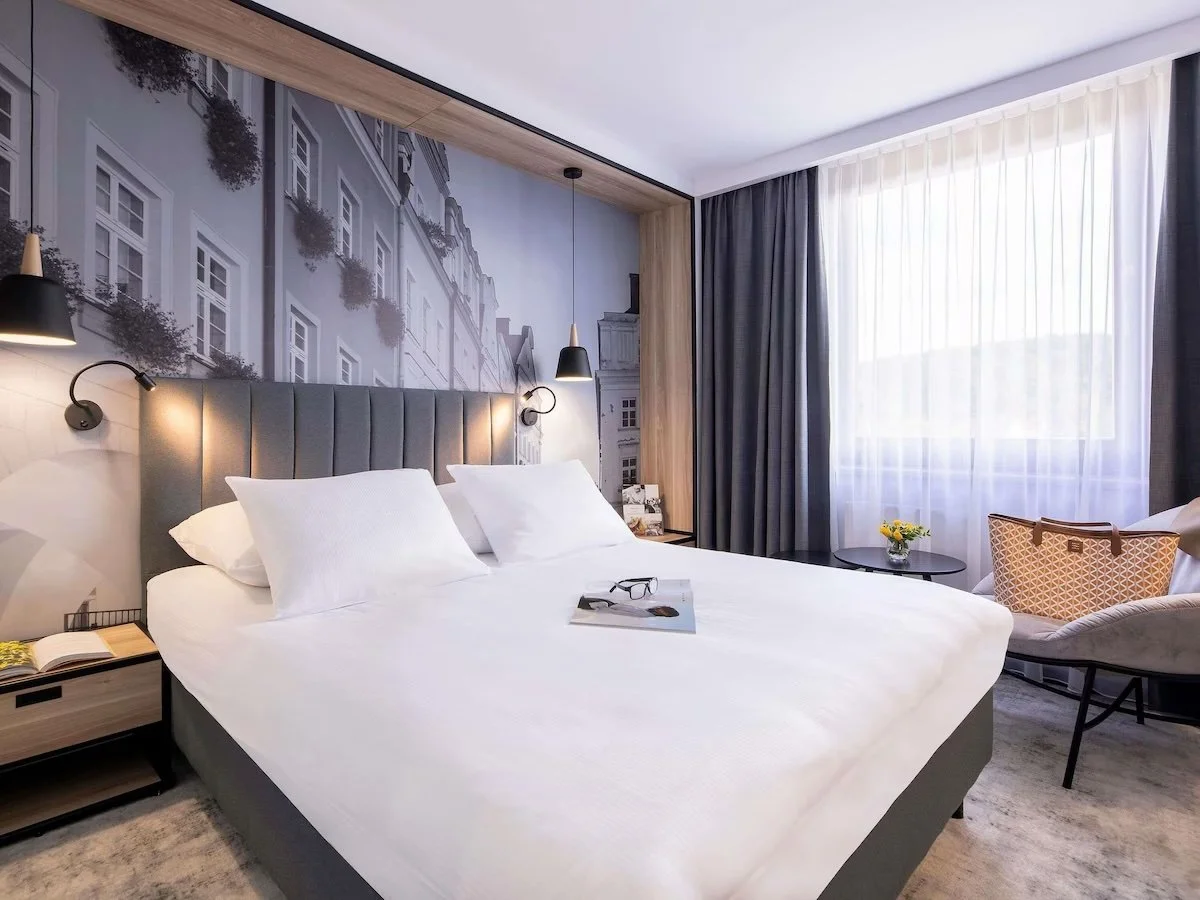 A modern hotel room with a large bed featuring white linens and pillows, a wall-mounted lamp on each side, a mural of city buildings behind the bed, a window with sheer white and dark curtains, a small round table with a vase of yellow flowers, and a