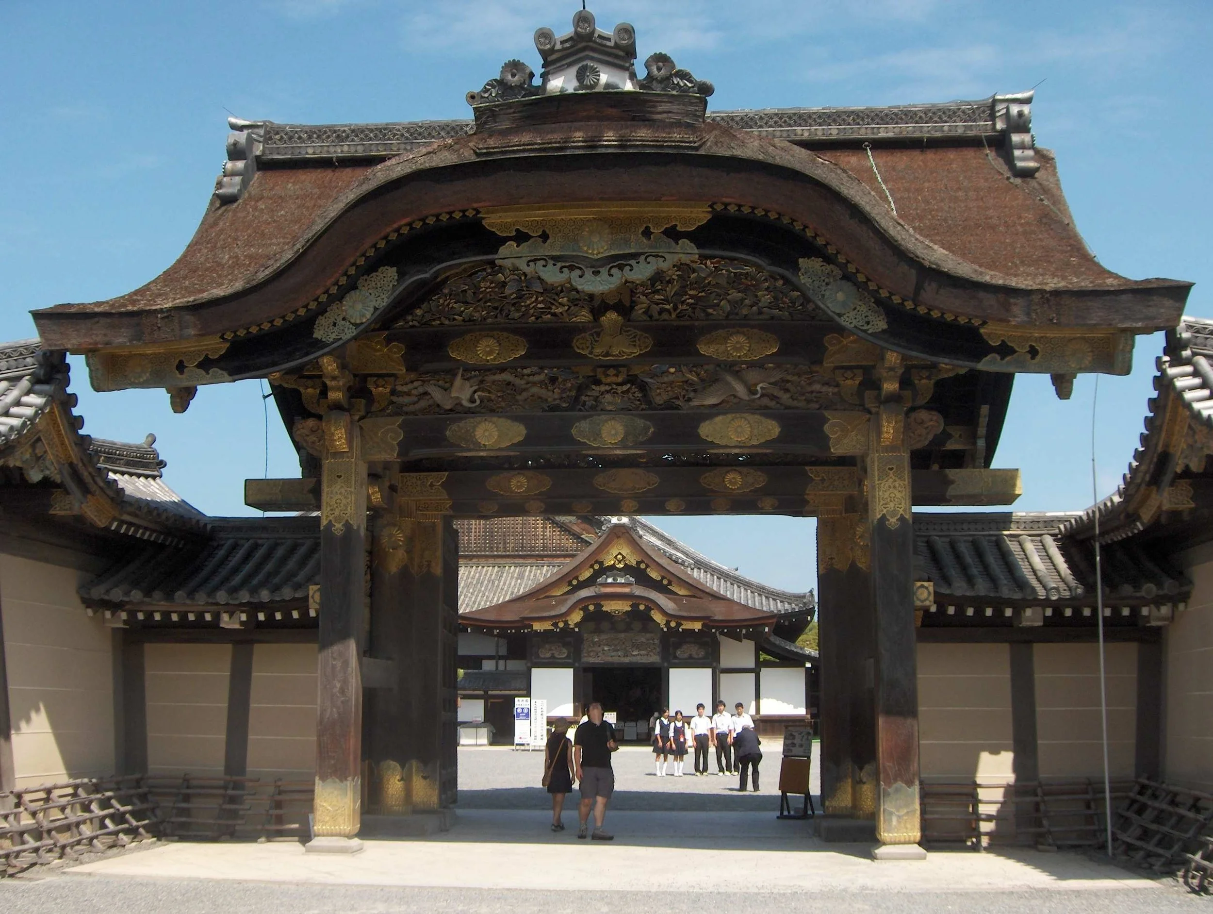 Traditional Japanese temple gate with intricate woodwork and gold accents, and a group of people walking underneath.
