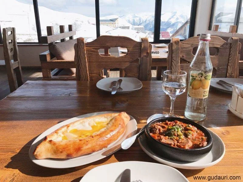 A wooden dining table with a plate of khachapuri, a bowl of stew, a glass of water, a bottle of flavored water, and a set of utensils, with a mountain view in the background.