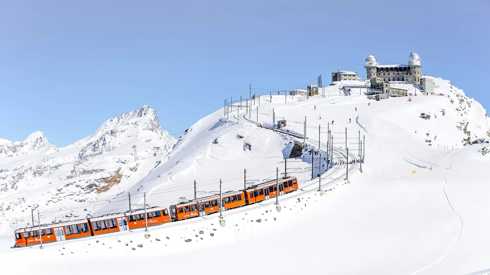 An orange train traveling through snow-covered mountains toward a building with observatories on a ridge under a blue sky.