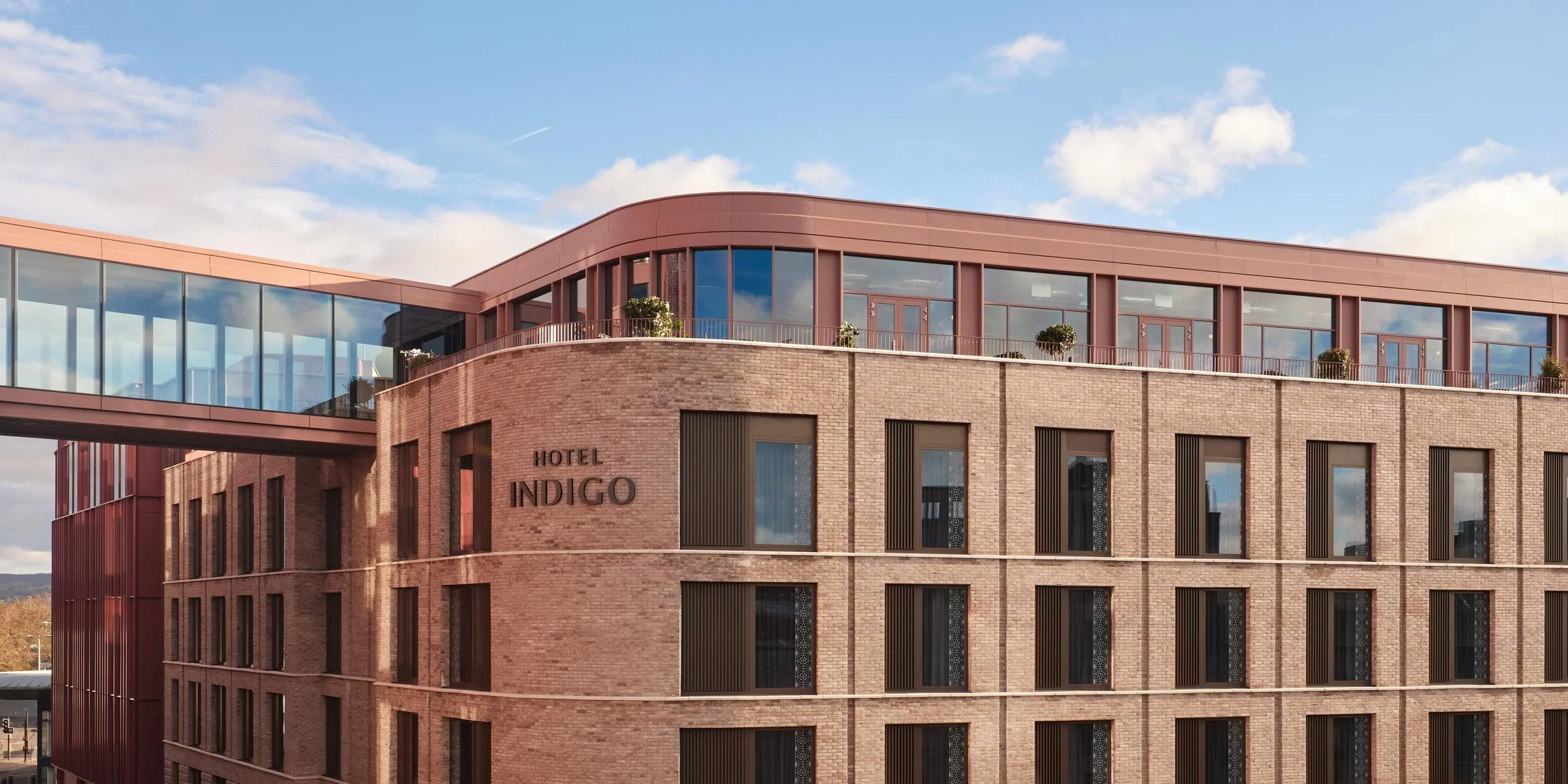 Exterior view of Hotel Indigo with brick facade, large windows, and rooftop terrace with potted plants, under a partly cloudy sky.