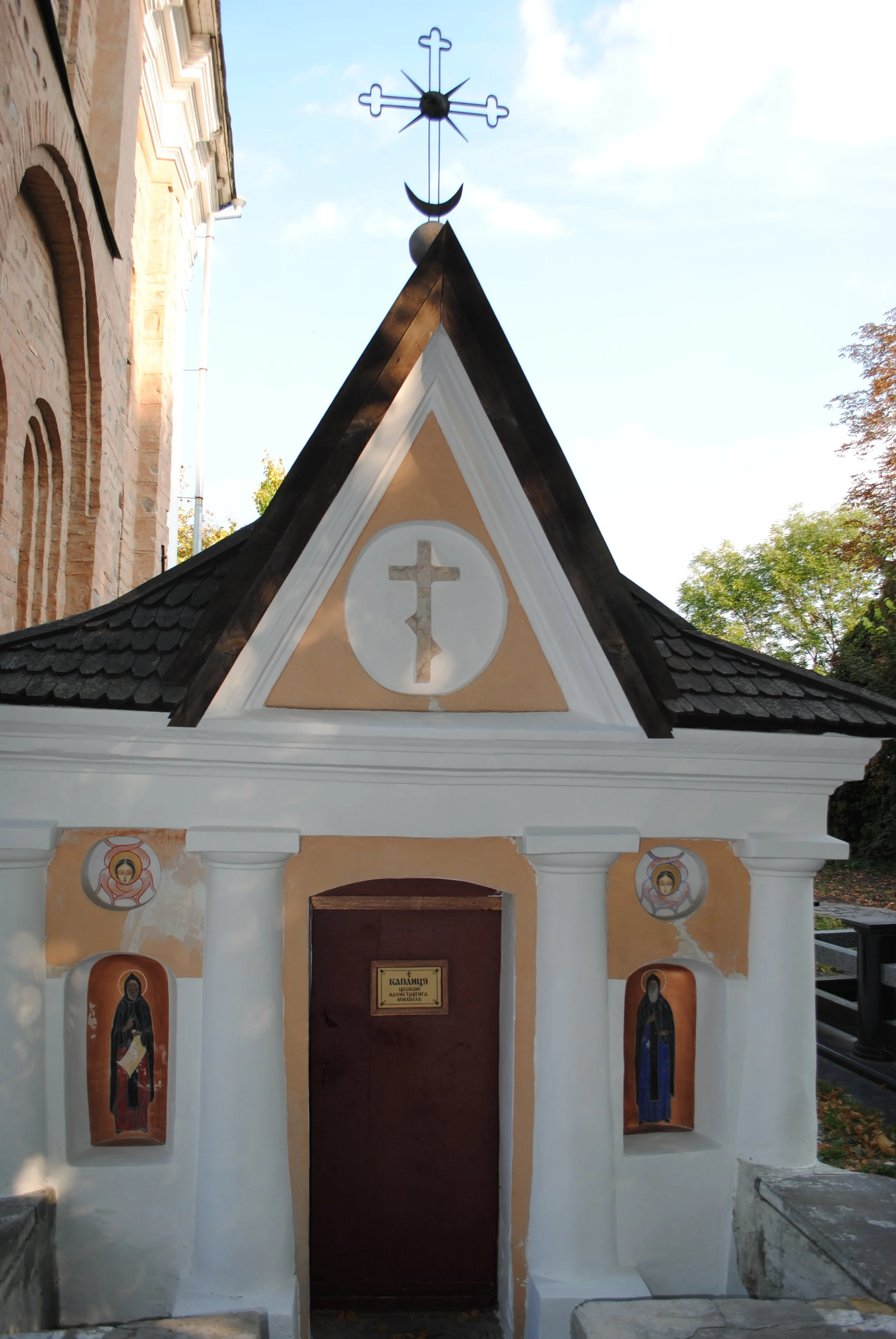 Small church or chapel with a triangular roof, featuring a cross and a crescent moon on top. The front has religious icons painted above the entrance, with four painted portraits of saints or religious figures below, set within arched niches, and a p
