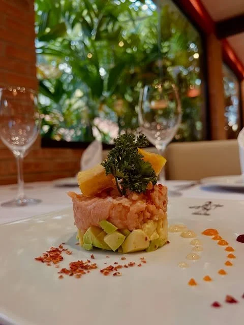 A plated dish featuring a layered salad or appetizer with diced avocado, a slice of pineapple, a dollop of pinkish meat or seafood, garnished with parsley, and arranged with decorative sauce and spice patterns on a white plate, set in a restaurant wi