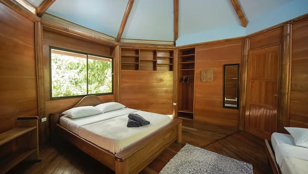 A bedroom with wooden paneling, a bed with white linens and pillows, dark towels folded on top, a window with green foliage outside, and built-in wooden shelves and closet.