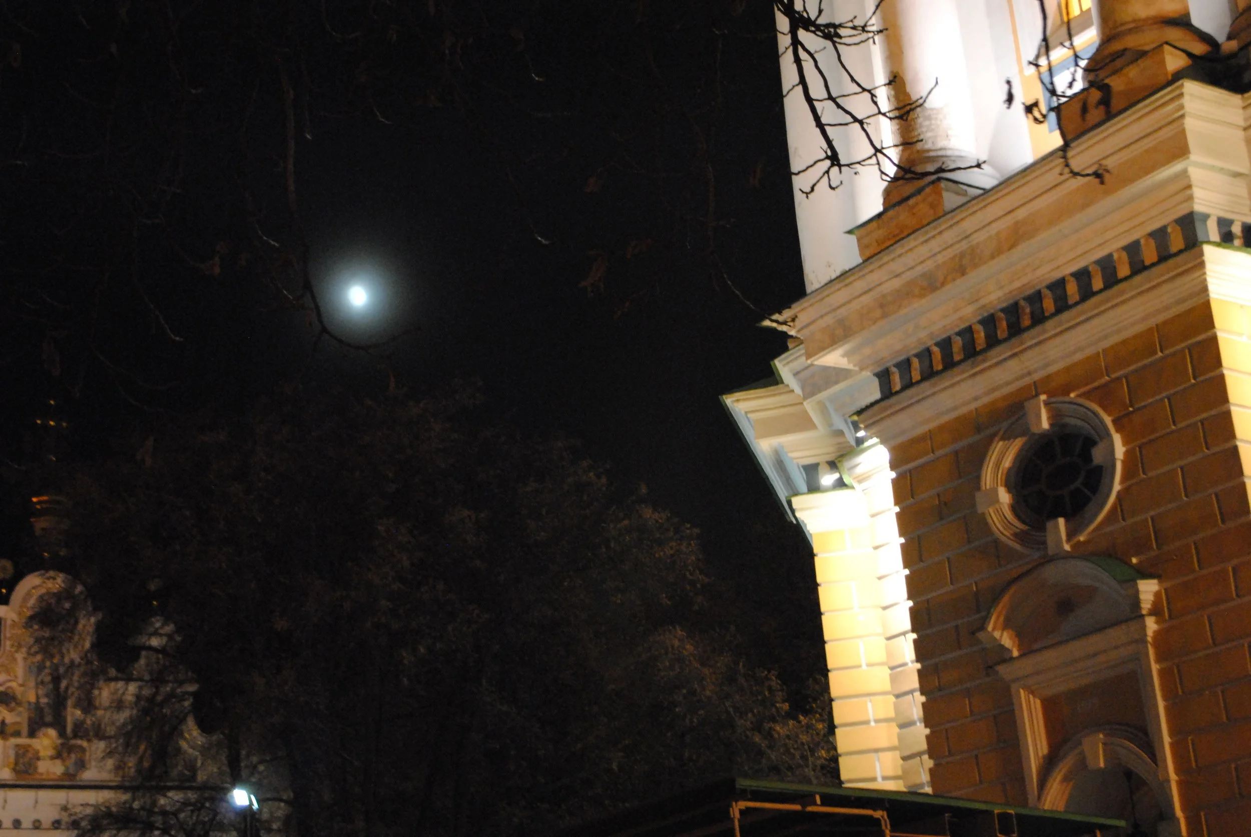 Nighttime view of a building with brick and white columns, illuminated by exterior lights. The sky is dark, with the glow of the moon visible behind bare tree branches.