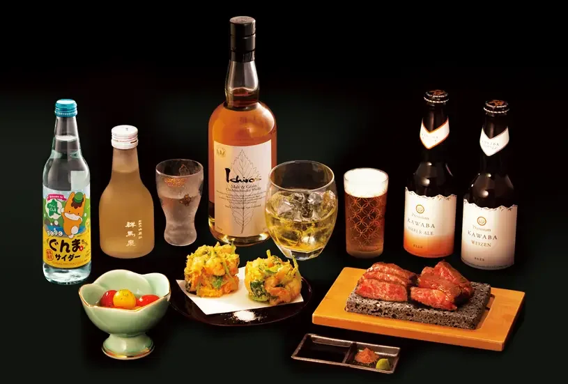 An assortment of beverages including sake, whiskey, wine, and beer, along with Japanese dishes such as grilled meat, tempura, a bowl of pickled vegetables, and small dishes with sauces on a black background.