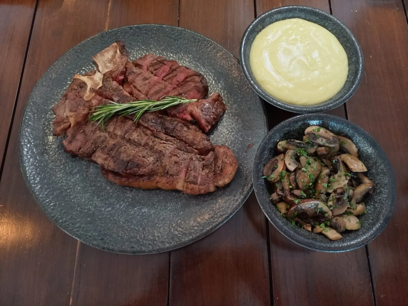 Grilled steak with rosemary on a dark gray plate, served with mashed potatoes and sautéed mushrooms garnished with chopped herbs, on a wooden table.