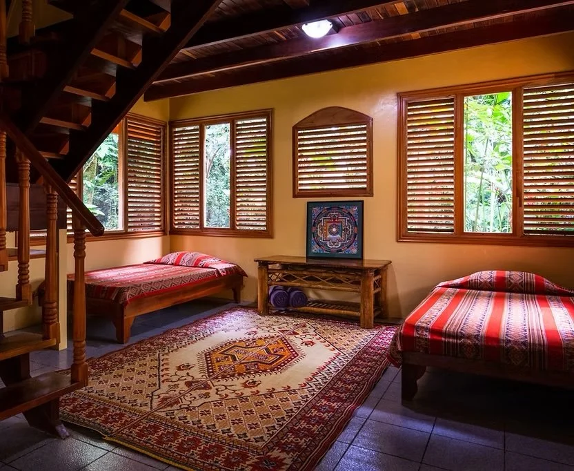 Interior of a rustic bedroom with two beds, wooden furniture, patterned rugs, and numerous windows with wooden shutters, surrounded by greenery.