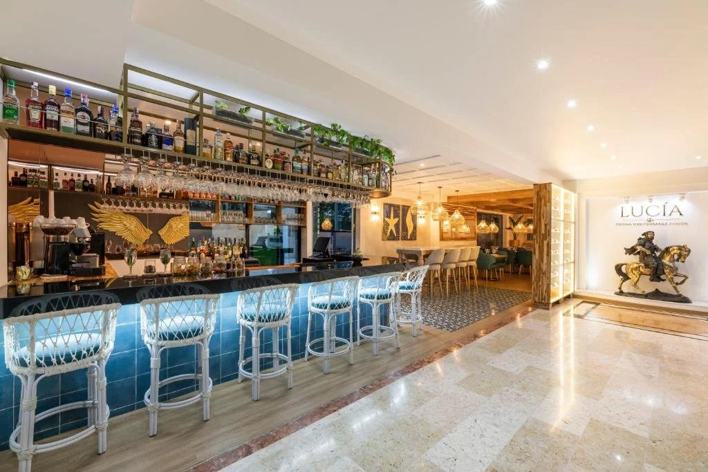 Interior of a modern bar and restaurant with a blue-lit bar counter, bar stools, and seating area with tables and chairs, decorated with artwork and sculptures.