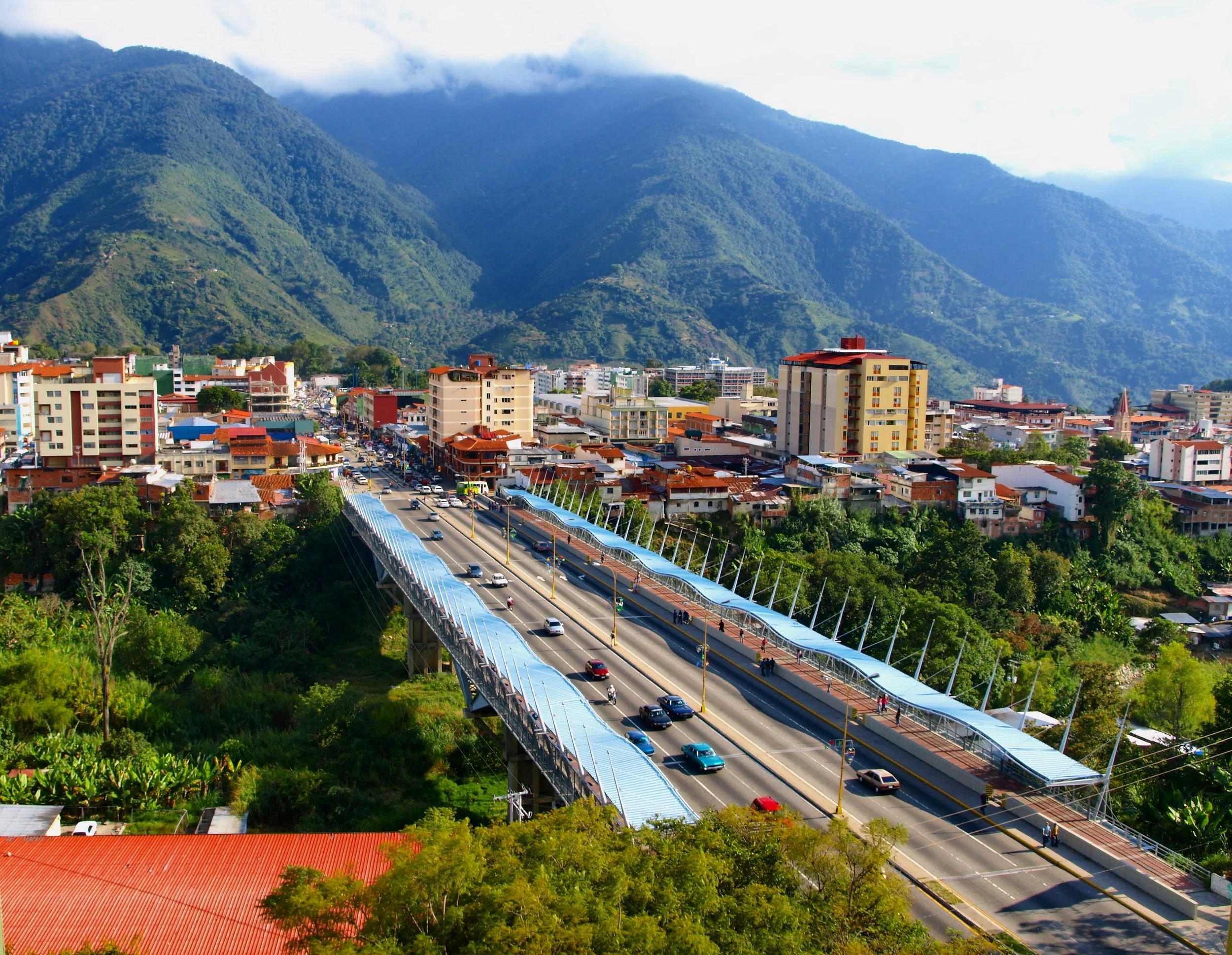 Cityscape with buildings and a bridge, set against green mountains and cloudy sky.