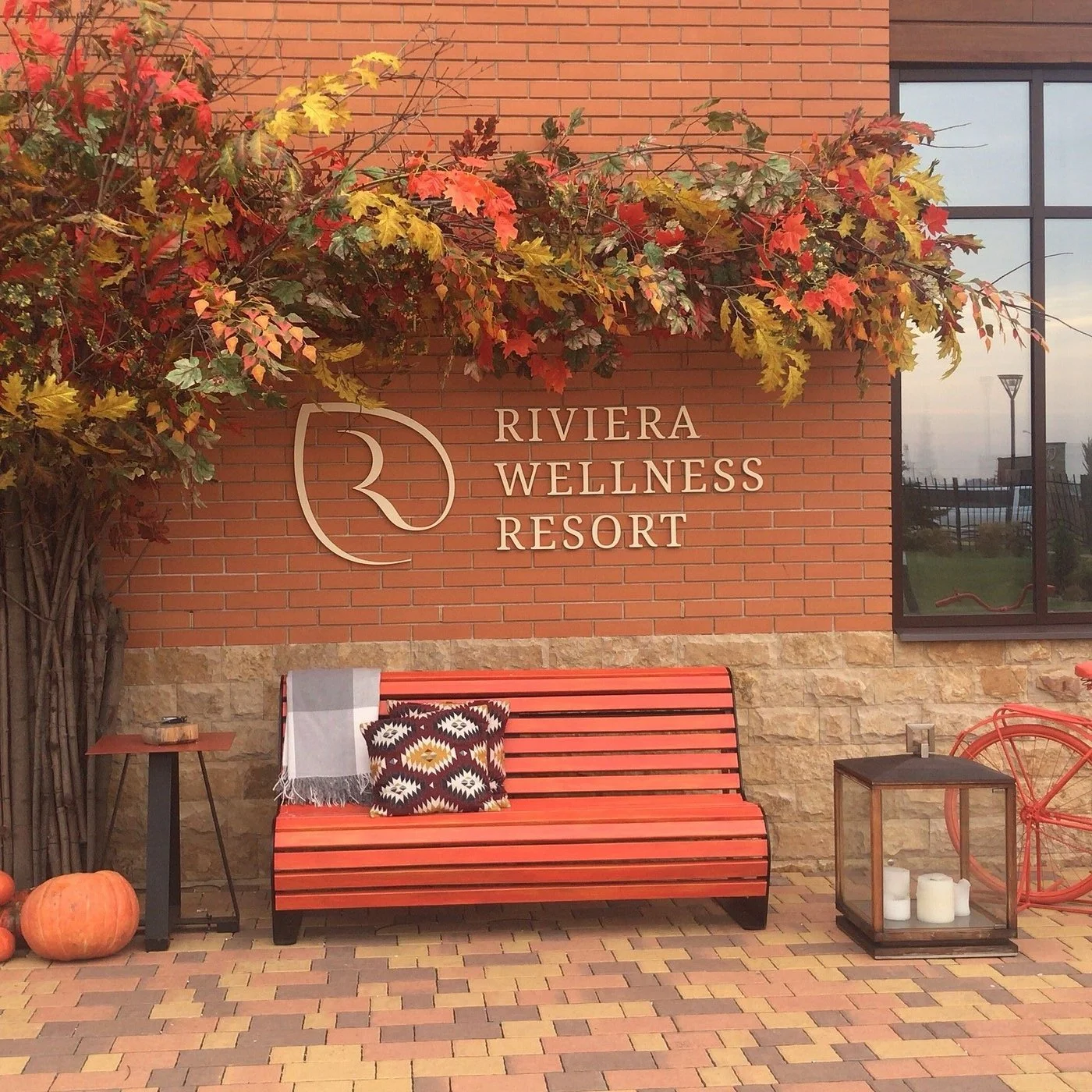 Exterior of Riviera Wellness Resort with a brick wall, large window, bench with pillow and blanket, pumpkins, and autumn leaves.