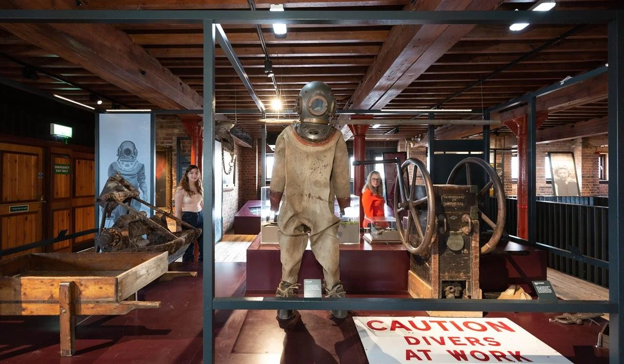 Museum exhibit featuring vintage deep-sea diving suit, gears, and machinery, with two women observing displayed artifacts.