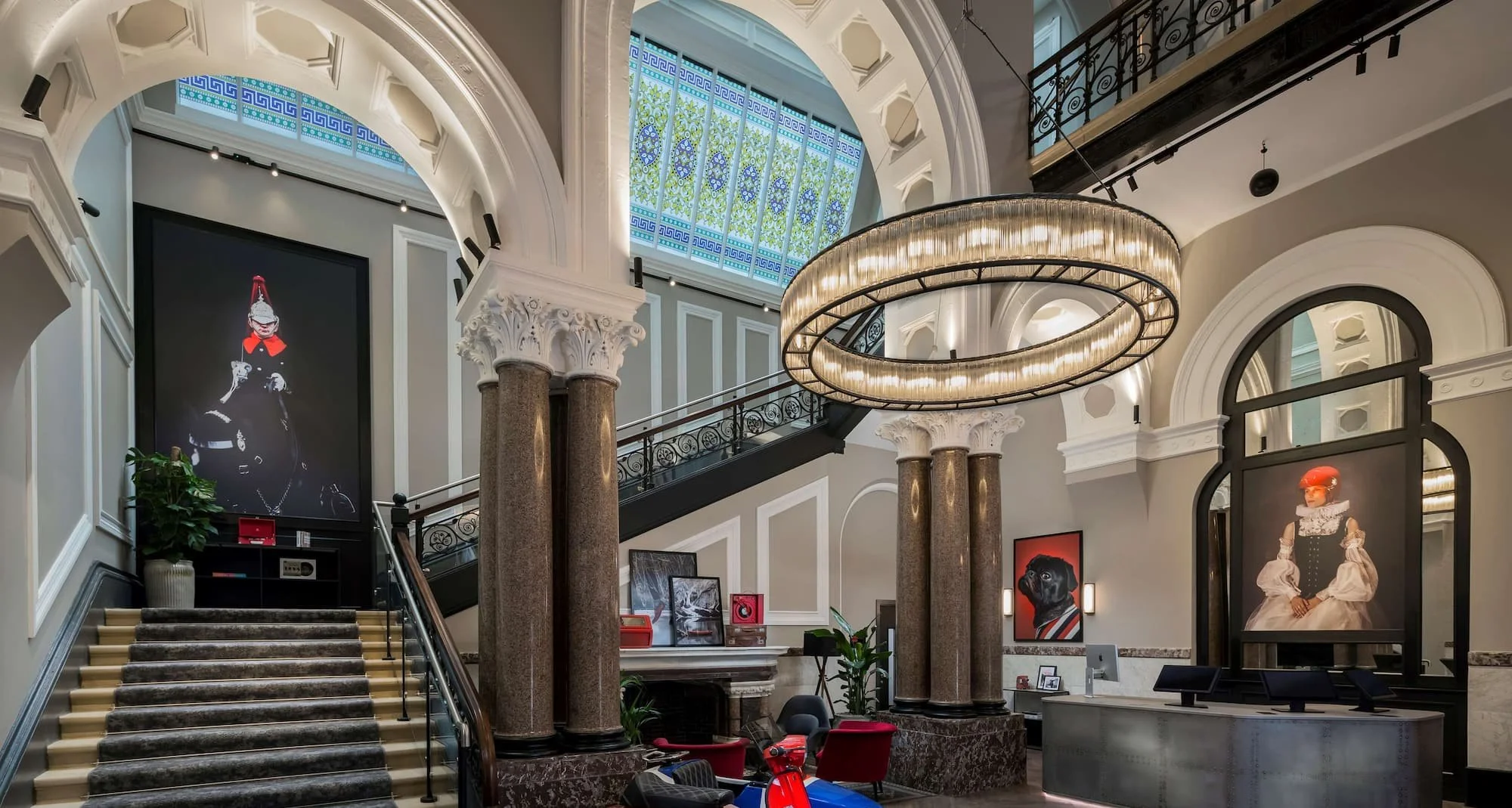 Interior of a grand building with a staircase, large decorative chandelier, and artwork of dogs and fashion models displayed on the walls.