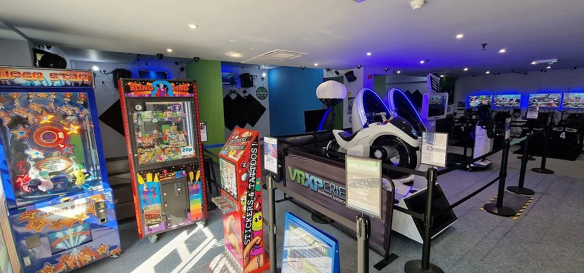 An arcade gaming area featuring claw and ticket machines, a VRXCR original motorcycle simulator, and multiple screens displaying racing games, all within a dimly lit space with black barriers and a colorful, modern design.