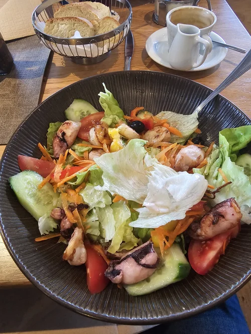 A large black bowl of mixed salad with lettuce, cucumbers, tomatoes, shredded carrots, shredded cheese, and grilled octopus slices.