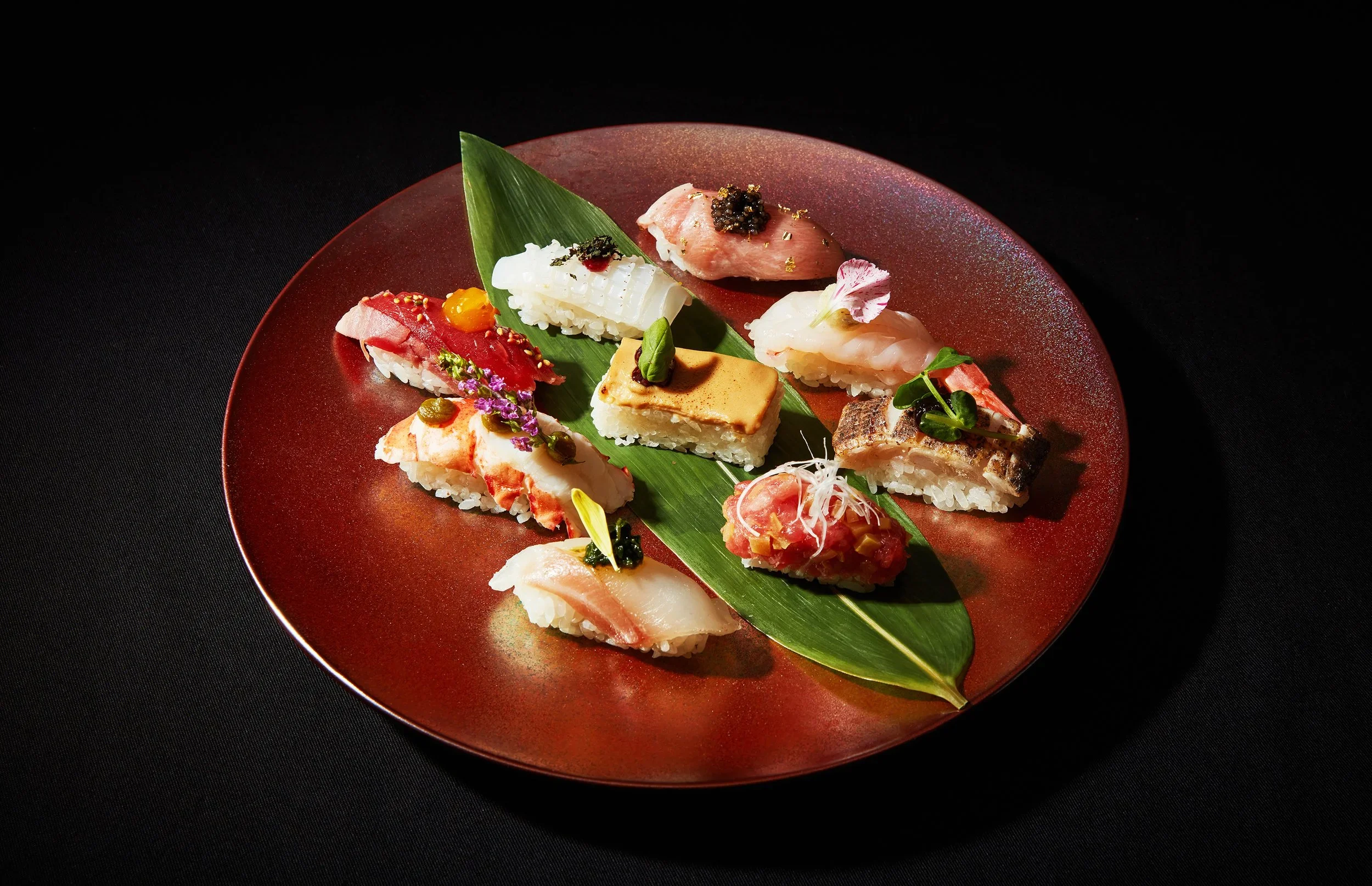 Assorted sushi pieces on a red plate with a green leaf garnish against a black background.