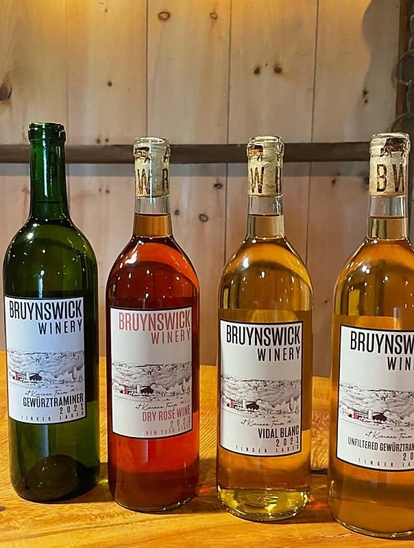 Four bottles of wine from Bruynswick Winery, displayed on a wooden surface. The bottles are green, red, white, and light amber, each with wine labels featuring mountain and vineyard imagery.