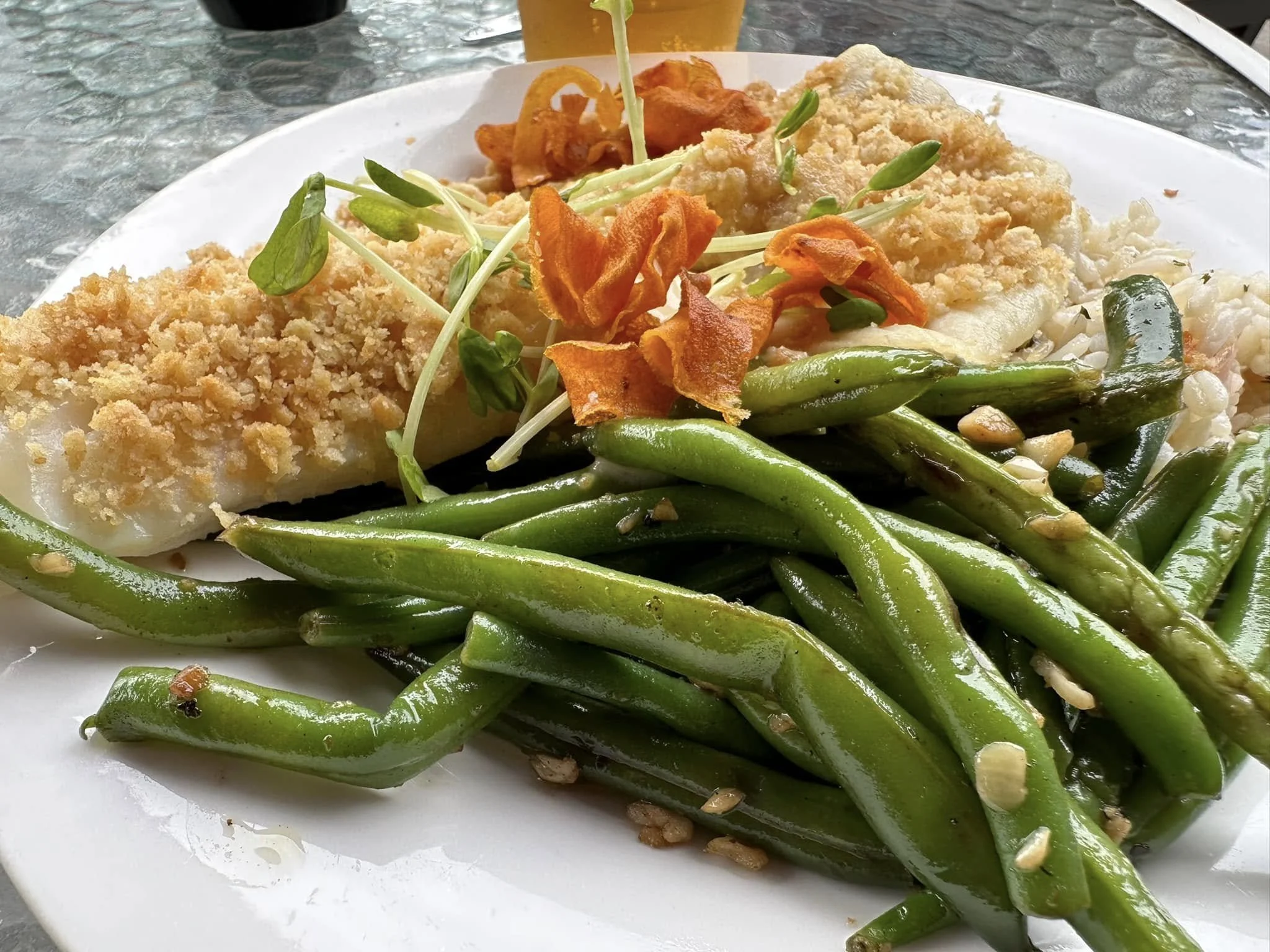 A white plate with green beans, topped with crushed nuts, on a glass table with a beverage in the background.