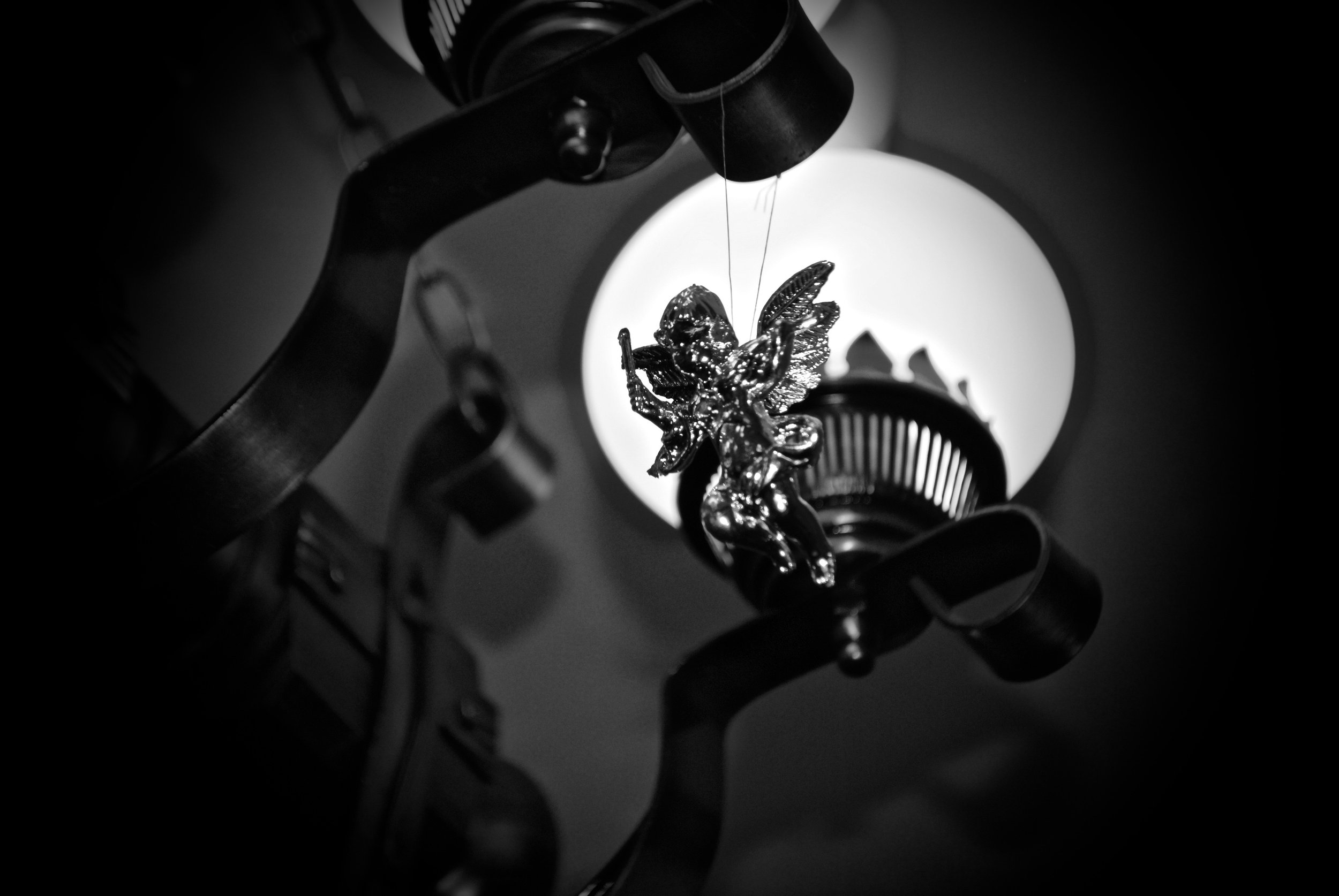 A black and white photograph of a spinning fairy or angel ornament hanging from a mechanical structure with wires, with a bright light source behind it creating a glowing effect.