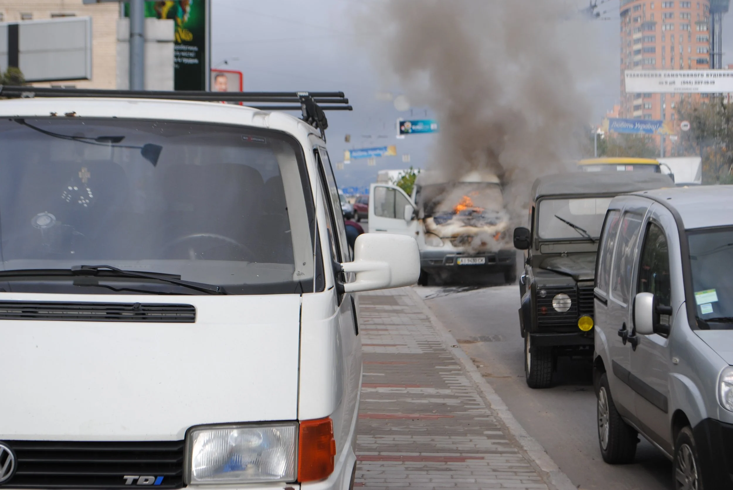 A street scene with a white van in the foreground, multiple parked cars, and smoke billowing from a vehicle with a visible fire in the background. Kiev November 2011, Marina Parrish ©