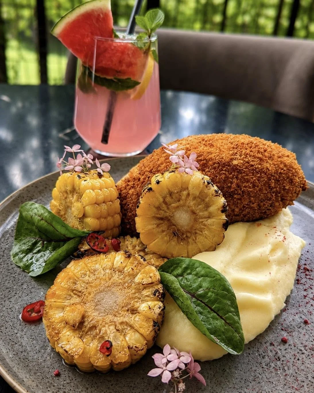 A plate with grilled corn, a fried croquette, and mashed potatoes garnished with herbs and flowers, with a pink fruit drink containing watermelon, lemon, and mint in the background.