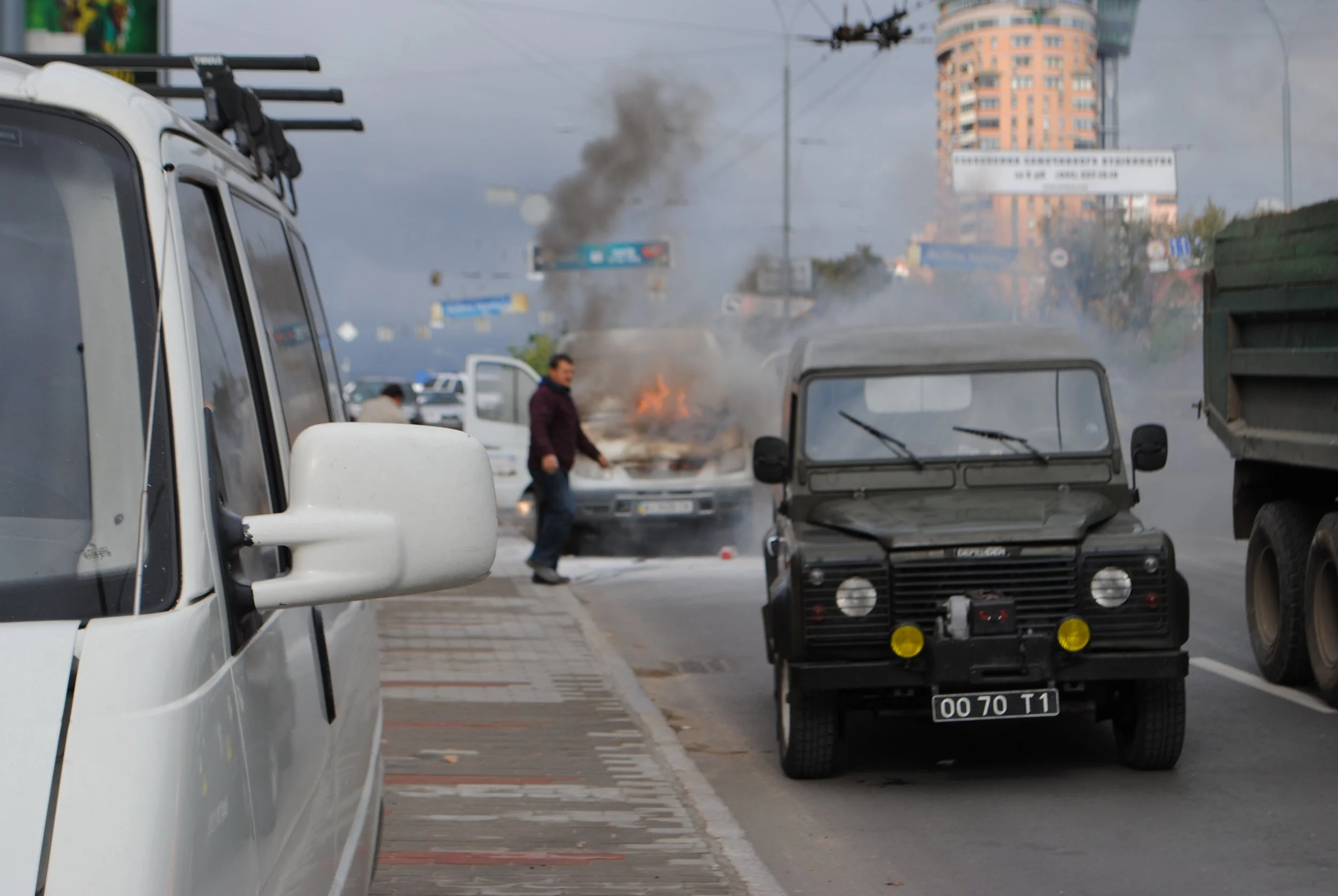 A street scene with cars parked along the curb, a vehicle on fire with black smoke rising, and people nearby, under a partly cloudy sky with a tall orange building in the background. Kiev November 2011, Marina Parrish ©