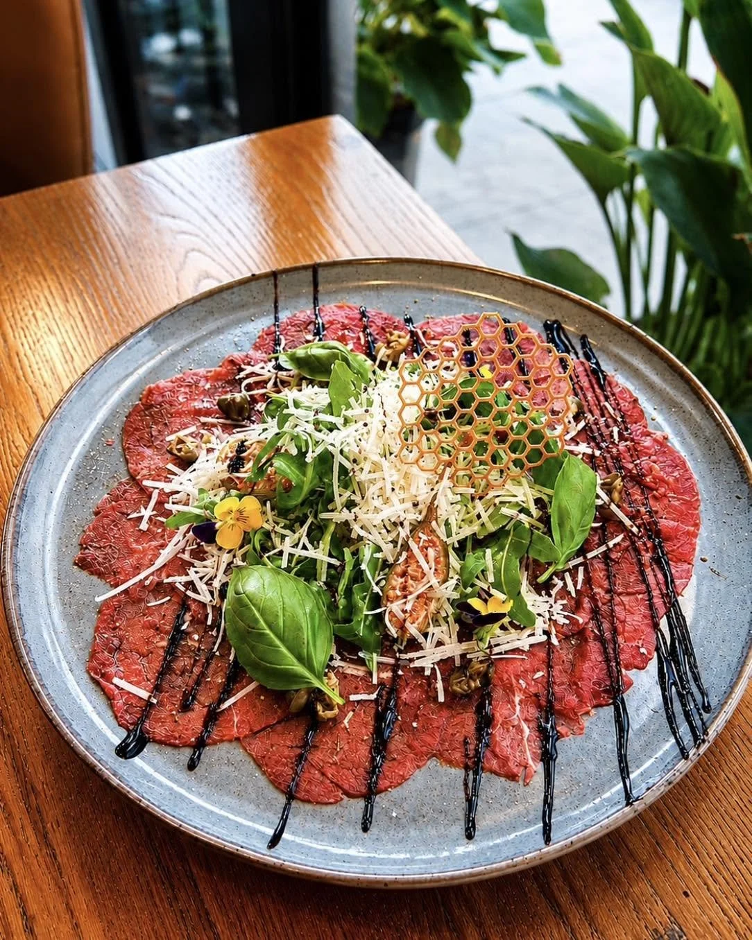 Plate of thinly sliced raw beef carpaccio topped with shredded cheese, basil leaves, microgreens, edible flowers, honeycomb piece, capers, and drizzled with balsamic and chocolate sauces.