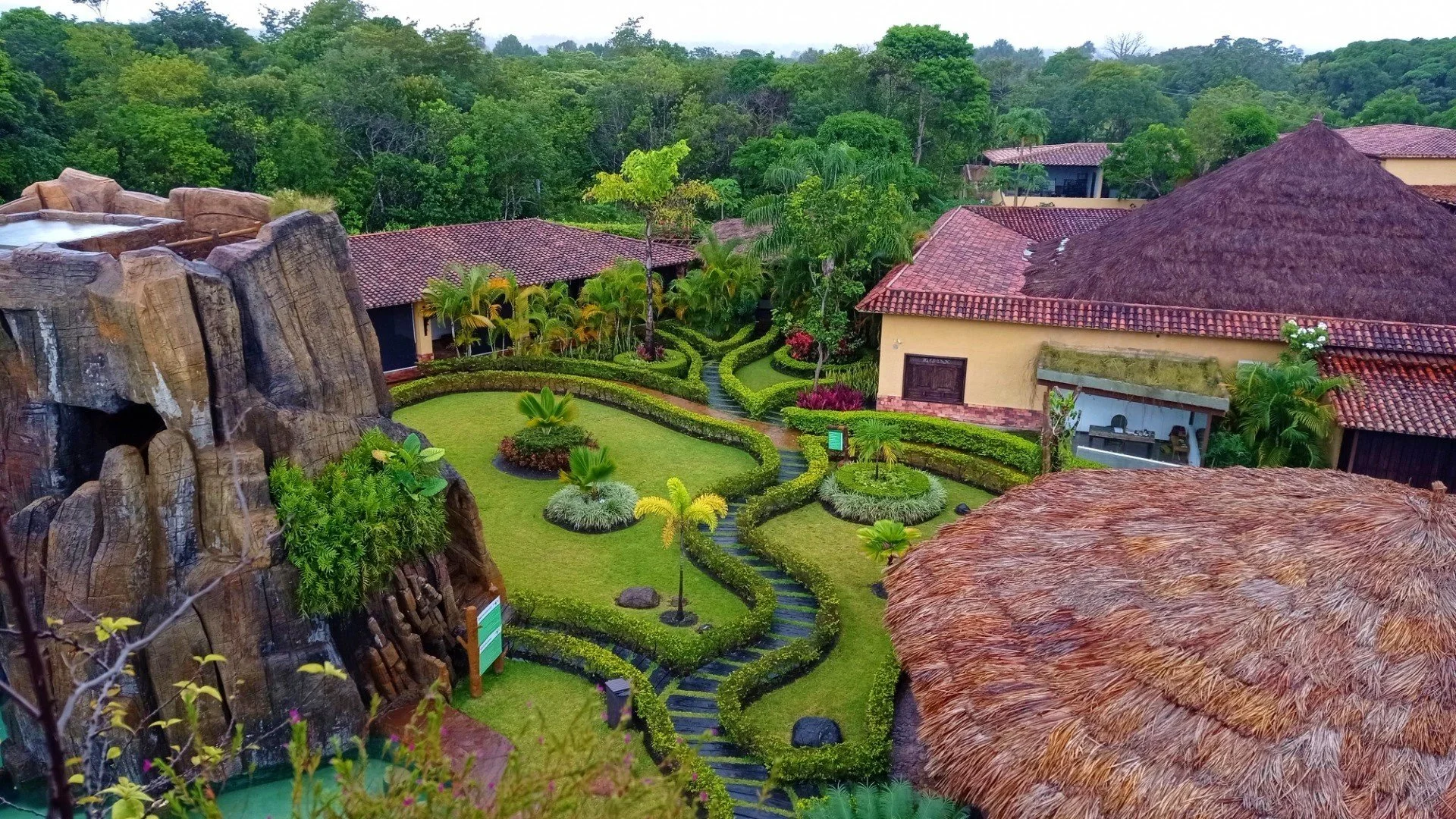 View of a lush garden with winding pathways, trimmed hedges, and small trees, surrounded by buildings with red-tiled roofs and thatched roofs, set in a tropical forest environment.