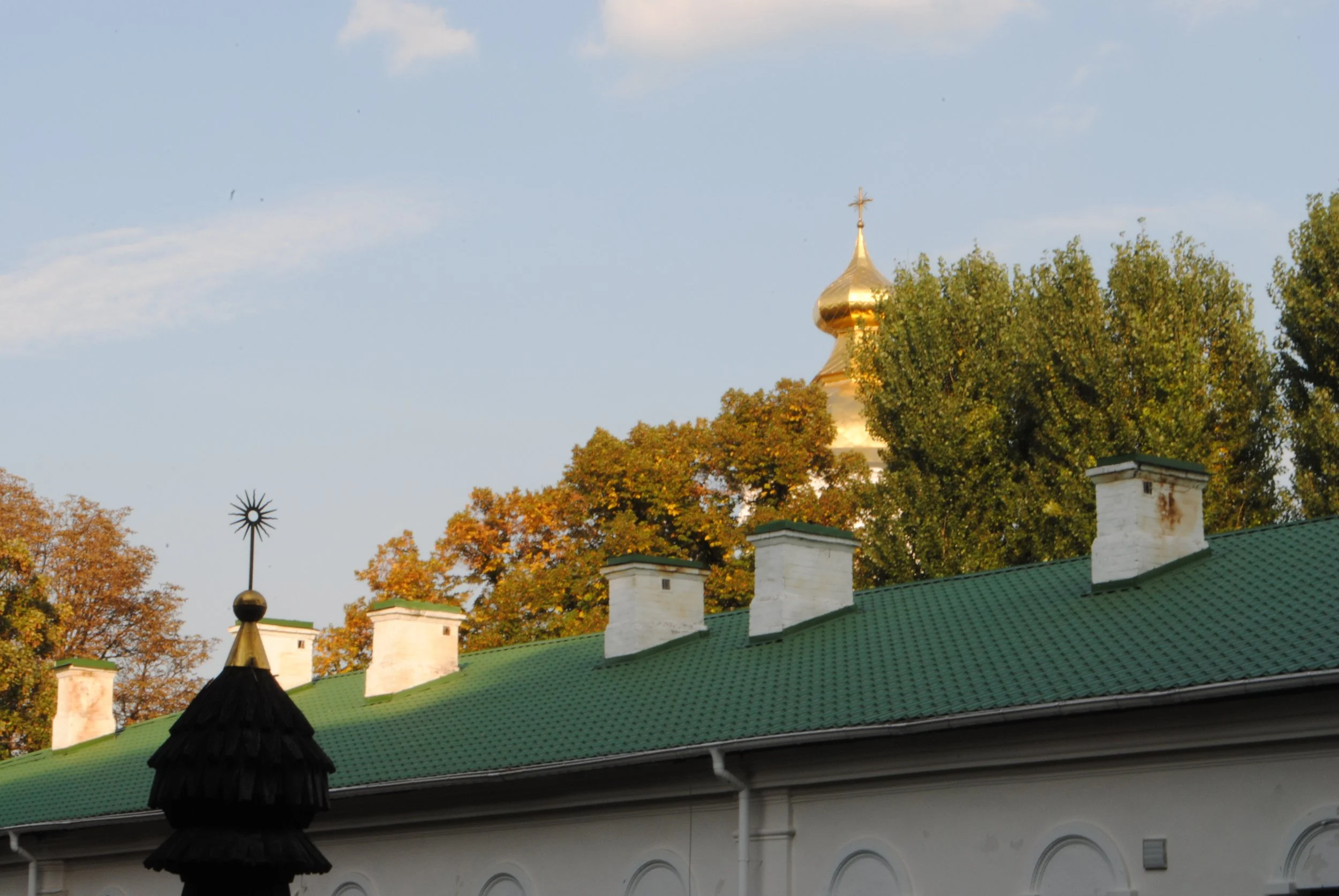 A building with a green roof and white walls, topped with white chimneys and decorative black and gold rooster weathervanes. In the background, a golden church dome and cross are visible above trees with autumn-colored leaves.