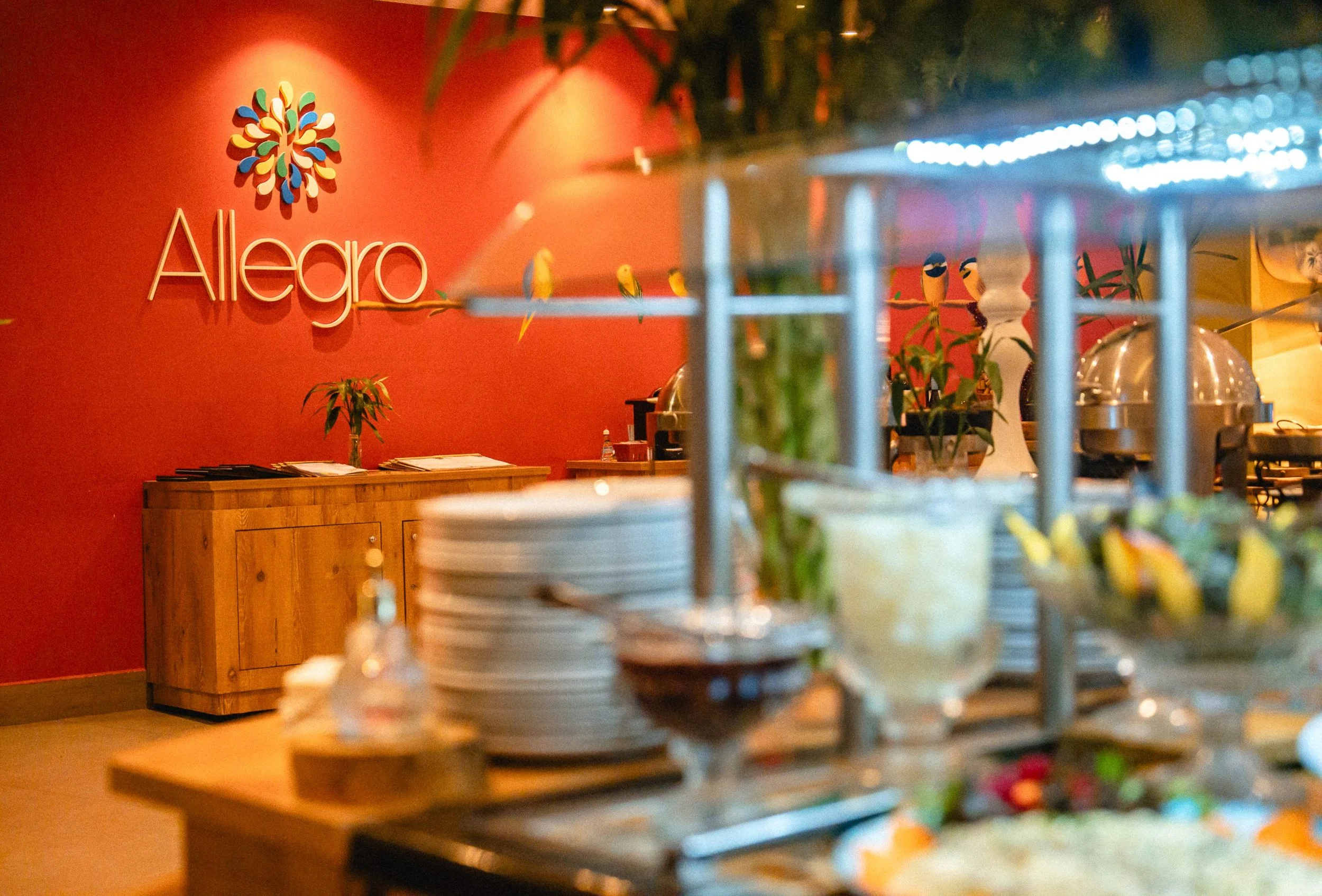 Interior of a restaurant with a red wall, featuring the logo 'Alegro' and a colorful tree-like design, with tables and dishes in the foreground.
