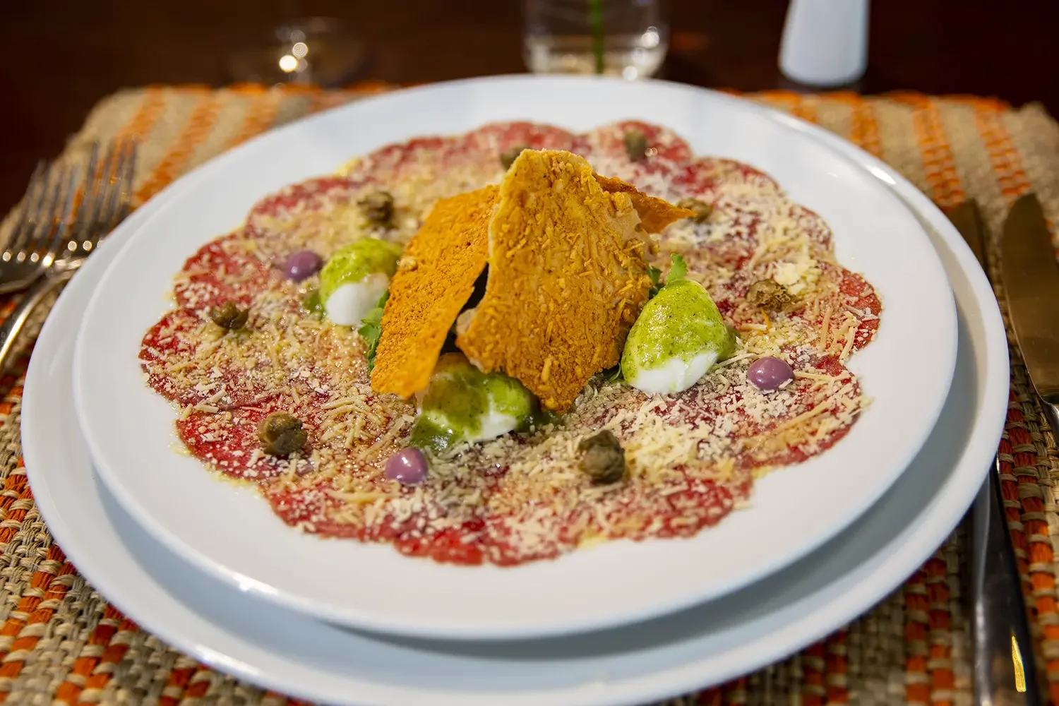 Plate of Italian dish, with beef carpaccio topped with cheese, capers, purple and green dollops of sauce, and crispy fried breaded shrimp in the center.
