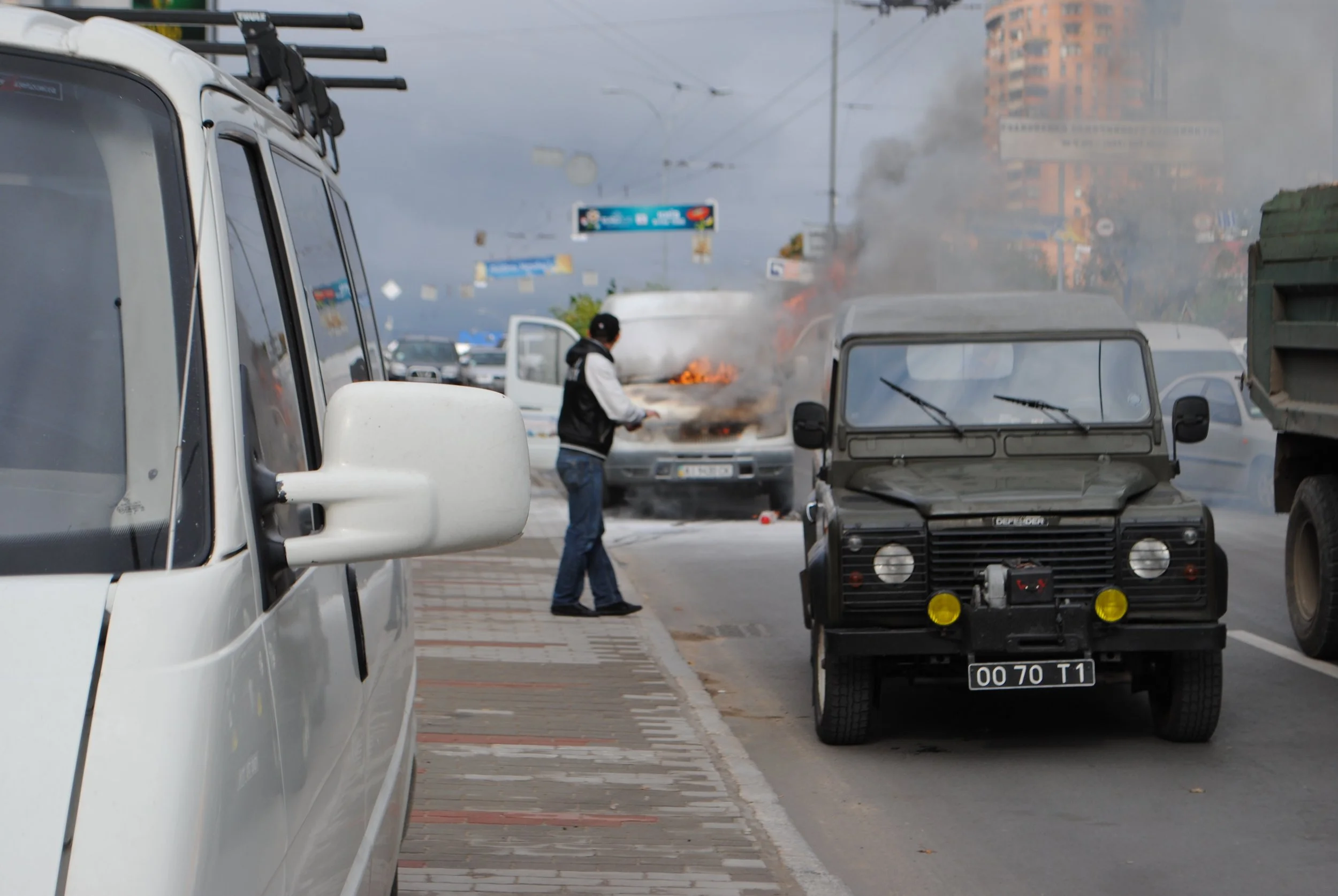 A street scene showing cars, with one vehicle on fire and thick black smoke billowing from it. A person is standing nearby, possibly trying to address the fire. Kiev November 2011, Marina Parrish ©