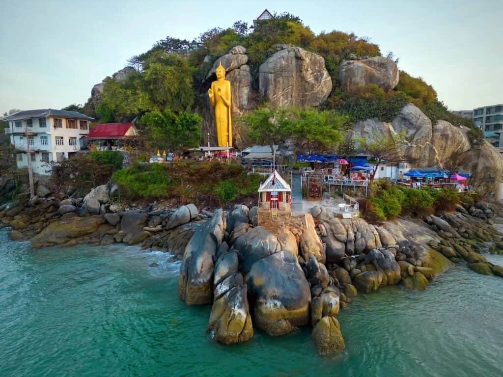A large golden Buddha statue on a rocky hill overlooking a seaside area with buildings and market stalls with umbrellas.