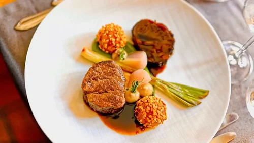 Appetizer plate featuring a steak medallion, roasted vegetables, and croquettes, garnished with herbs and sauces.