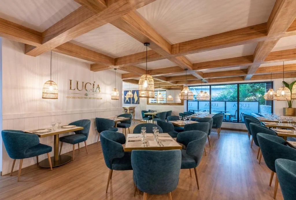 Interior of a modern restaurant with wooden ceiling beams, blue upholstered chairs around wooden tables, and pendant lighting fixtures, large window with outdoor view.