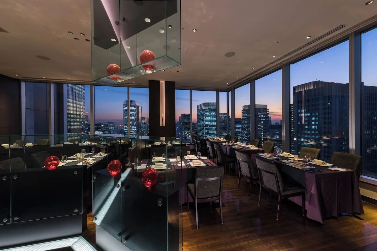 Interior of a high-rise restaurant or event space with large windows showing a city skyline at dusk, a long table set for dining with chairs, and decorative red glass balls hanging from the ceiling.
