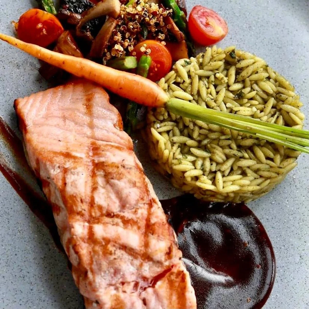 A plate with salmon fillet, cooked carrots, cherry tomatoes, green beans, a mound of orzo pasta, and a side of dark sauce.