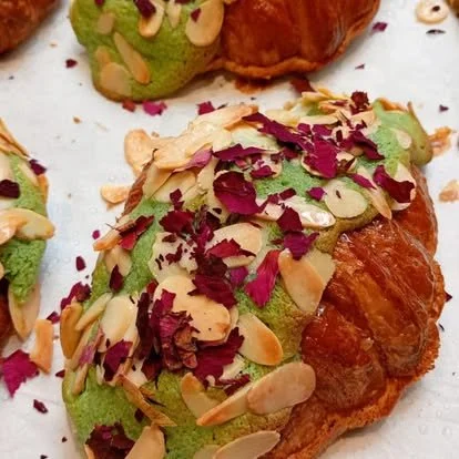 Close-up of a croissant topped with green frosting, sliced almonds, dried rose petals, and pistachios.