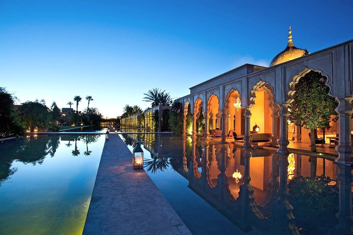 Elegant resort with a large reflecting pool, palm trees, and a classical building with arches and golden domes, illuminated at dusk.