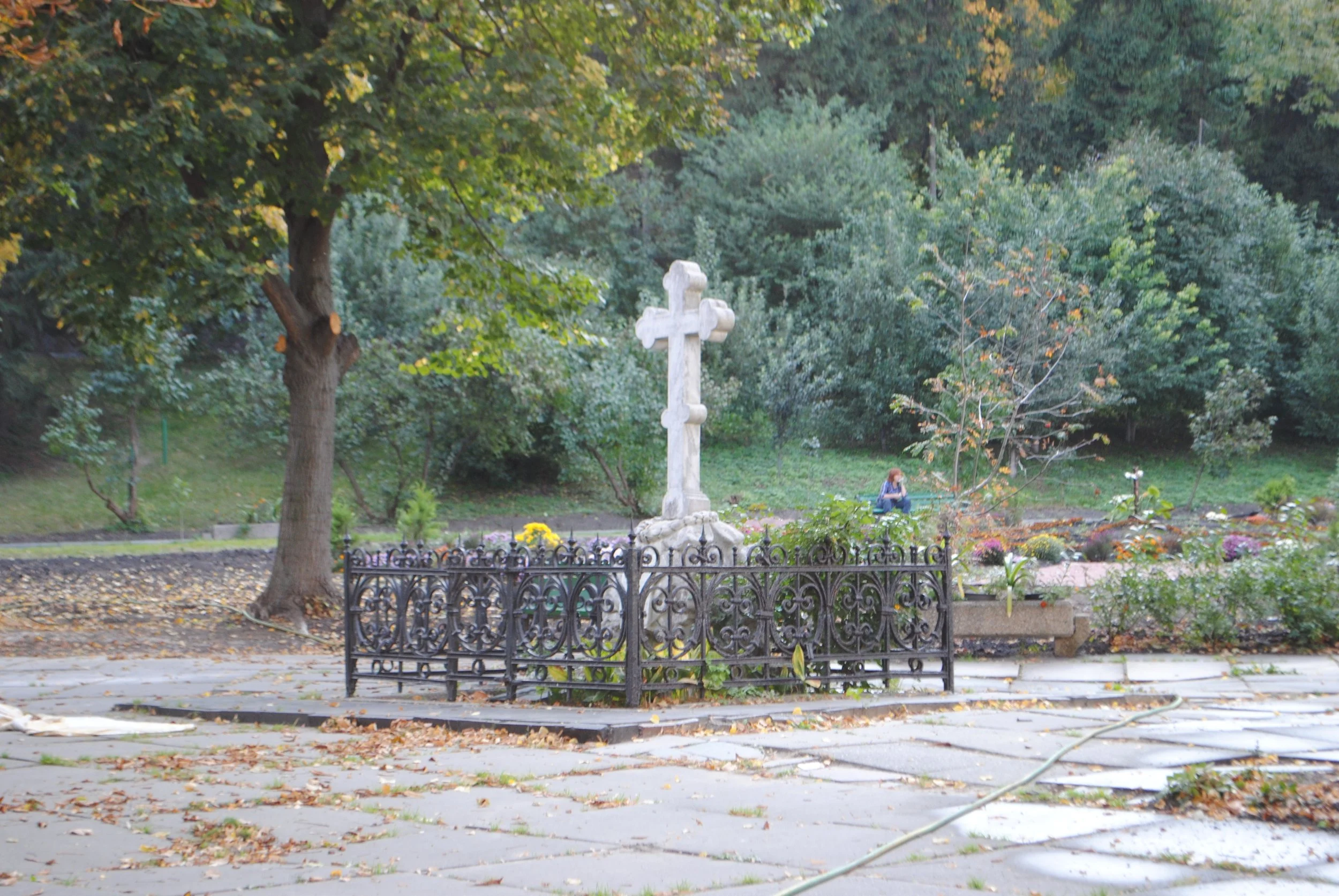 A white stone cross monument surrounded by a decorative black fence, set in a park with trees and a woman sitting on a bench in the background.