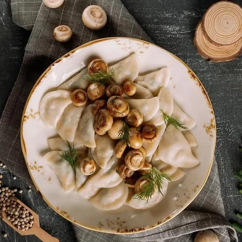 Plate of dumplings garnished with roasted mushrooms and fresh dill, served on a decorative plate, with wood and herbs in the background.