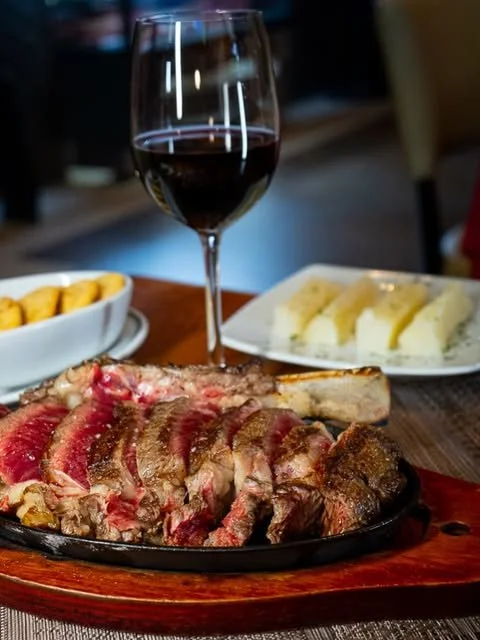 Steak served on a sizzling platter with a glass of red wine and side dishes of vegetables and potatoes on a wooden table.