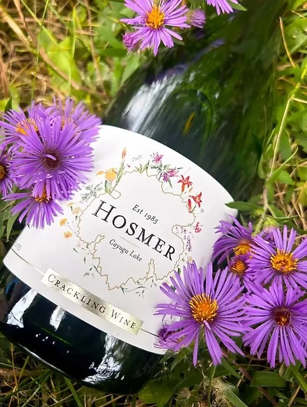 A bottle of red wine labeled 'Ho SmER' with 'Crackling Wine' written on it, surrounded by purple wildflowers lying on grass.
