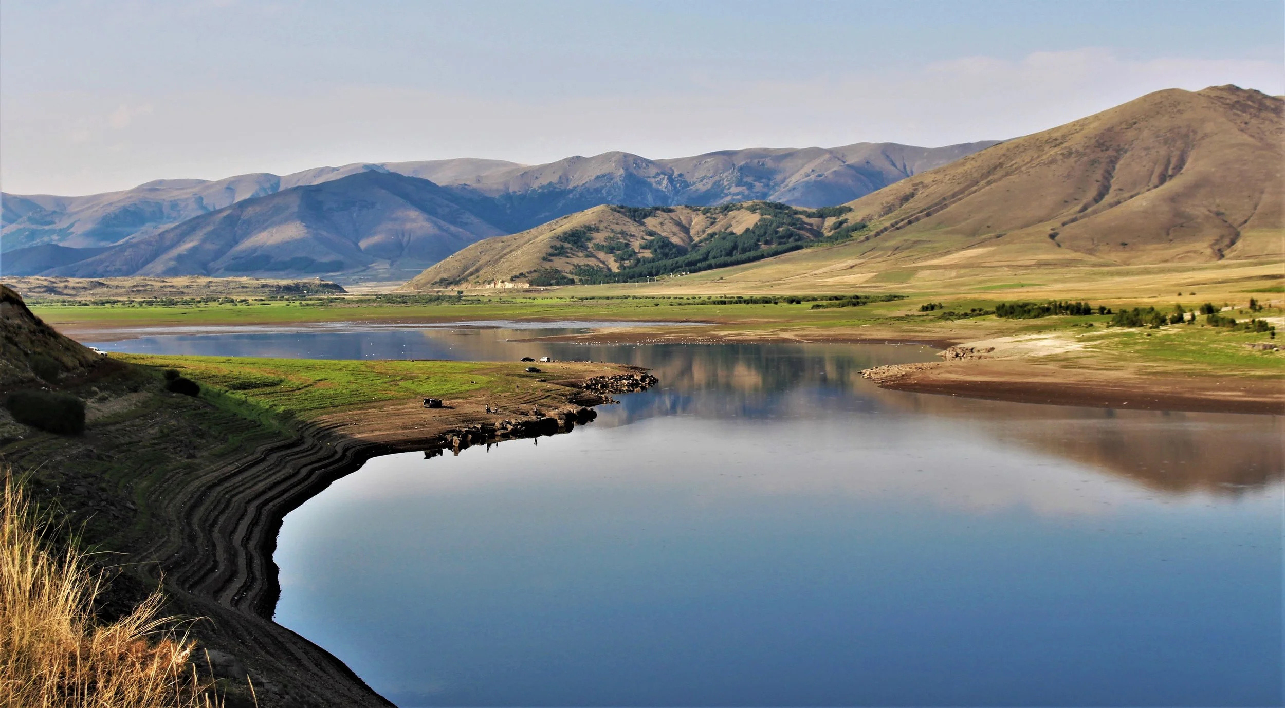 A landscape view of a river with mountains in the background and green fields on the riverbanks