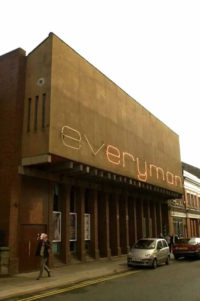 A building with a large neon sign displaying the word 'everymon' on its facade, with a few people walking by and cars parked on the street in front.