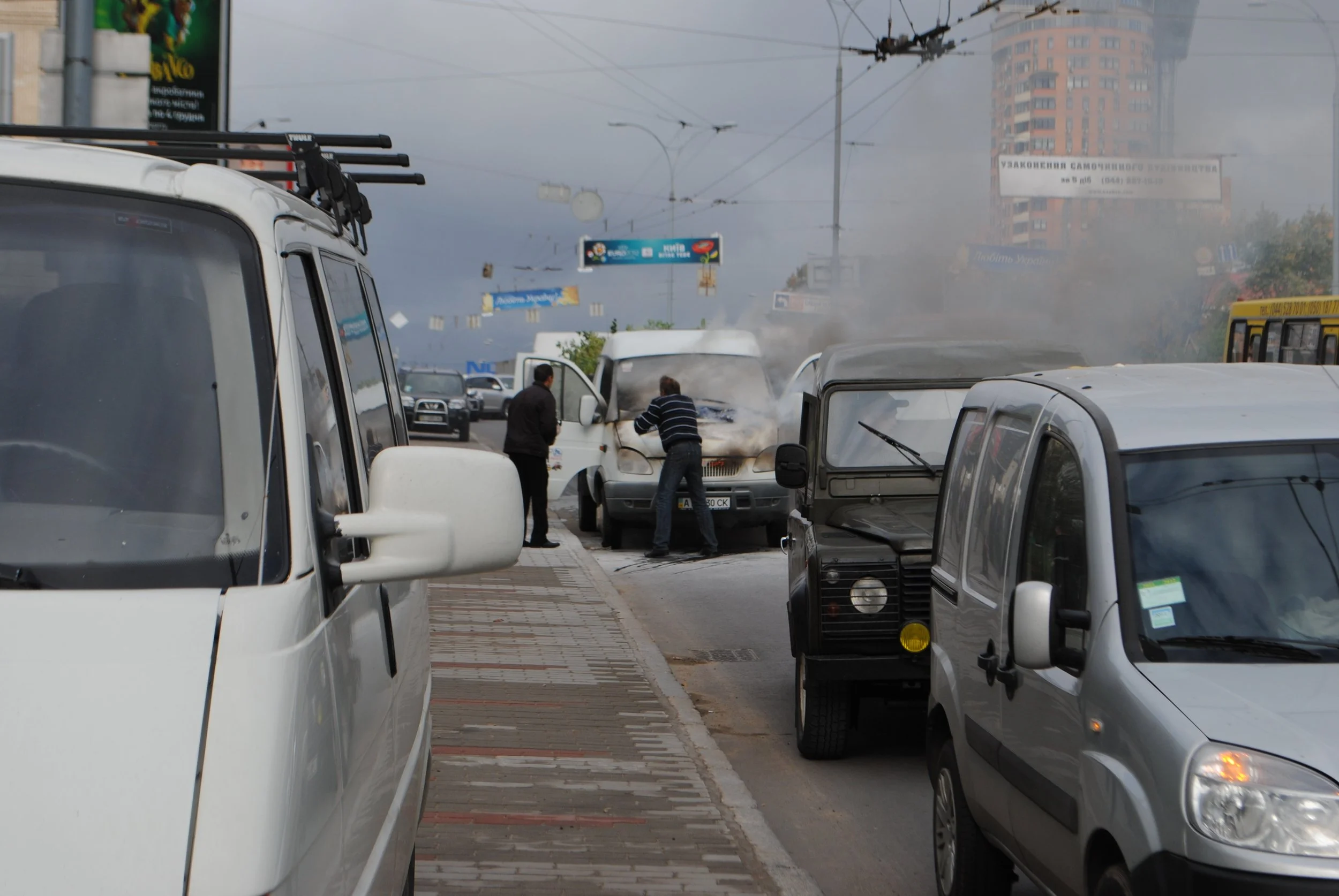 Two men inspect a car that is emitting smoke or steam from the engine on a busy city street, with several parked cars and a bus visible nearby and office buildings in the background. Kiev November 2011, Marina Parrish ©