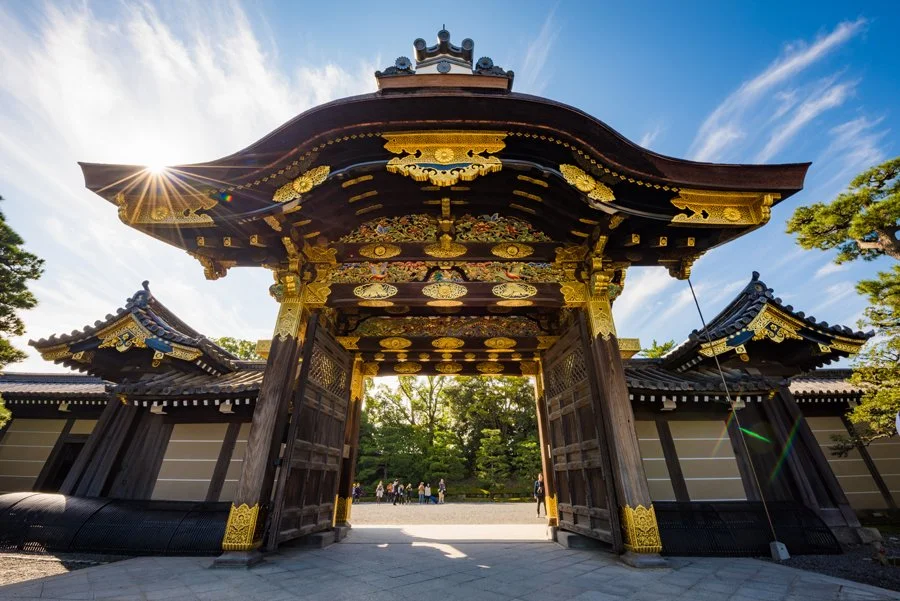 Traditional Japanese gate with ornate woodwork and gold accents, with a bright blue sky and trees in the background.