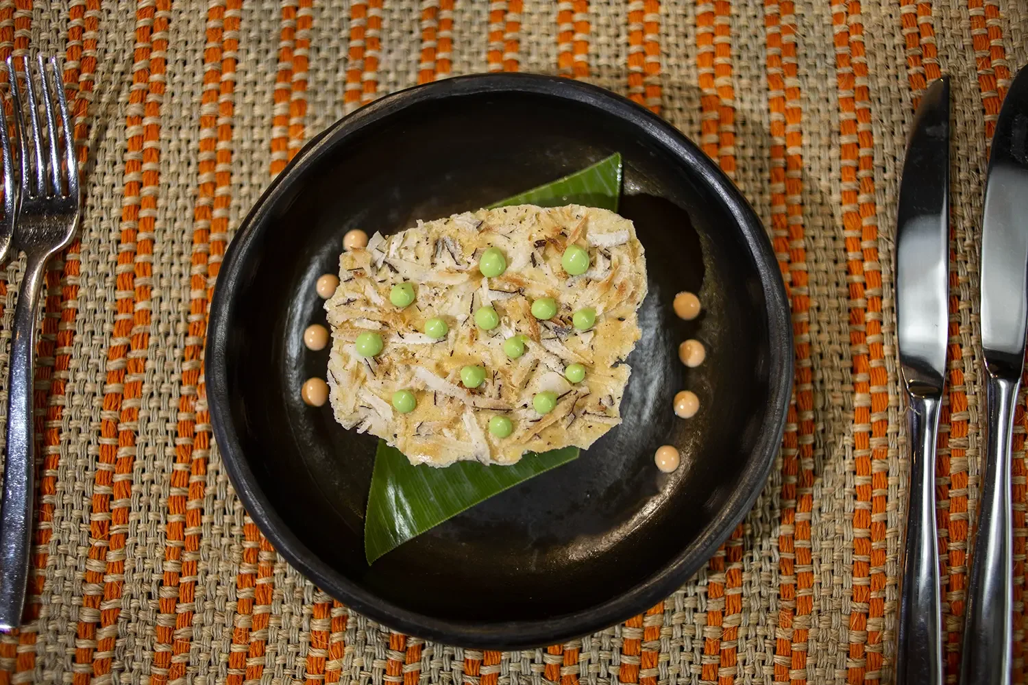 A piece of layered food topped with green and orange small round decoration, served on a black dish with a banana leaf underneath, on a multicolored woven placemat with silverware on the sides.