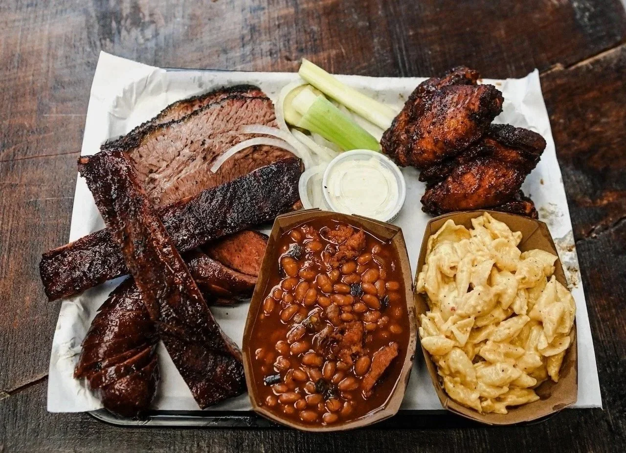 A tray of barbecue food including sliced beef brisket, smoked ribs, smoked sausage, baked beans, mac and cheese, and a side of sliced celery with a small container of sauce.