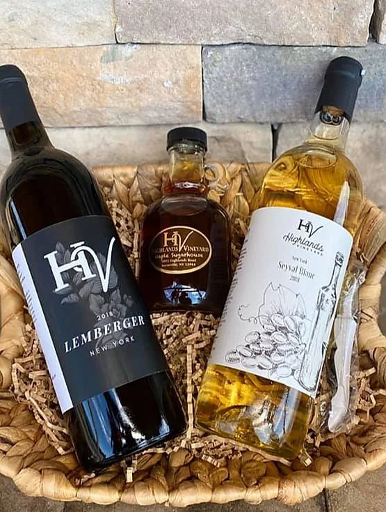 A gift basket containing a bottle of red wine, a bottle of white wine, and a small jar of syrup, all arranged on shredded paper in a woven basket against a stone background.